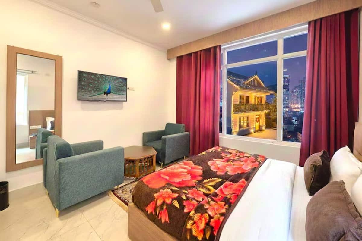 A hotel room with a large bed, two armchairs, a small table, a painting of a peacock, and a window with a city view at night.