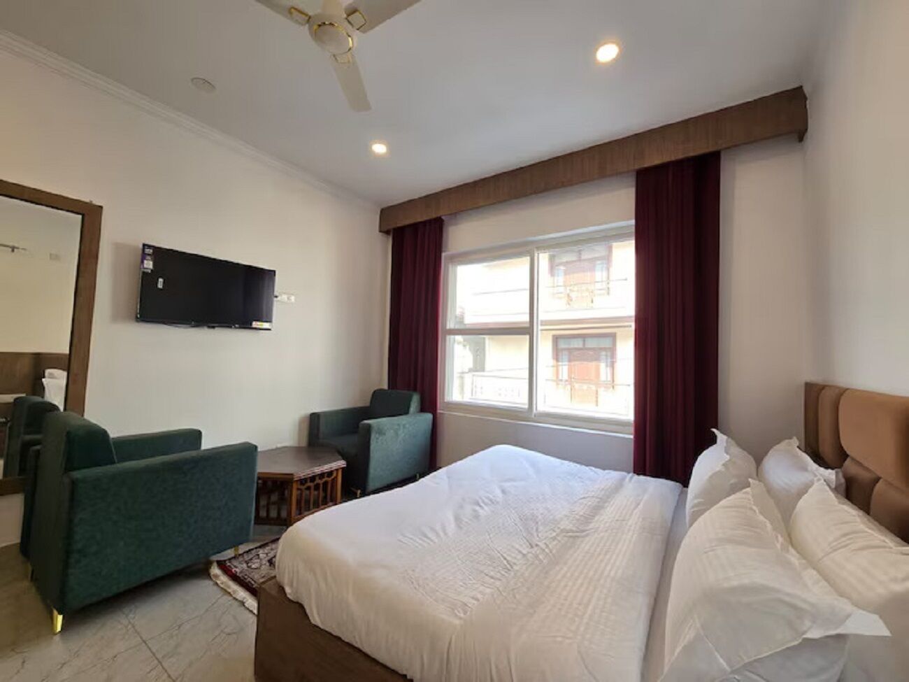 A hotel room with a large bed, two armchairs, a small table, a TV mounted on the wall, and a window with curtains.