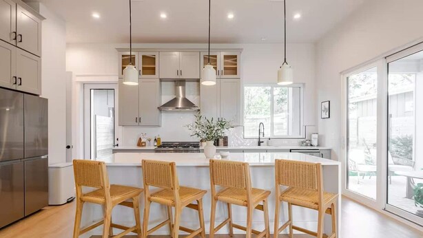 Een moderne keuken met een centraal eiland, vier rieten stoelen, roestvrijstalen apparatuur en grote ramen.