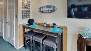 A dining area with a wooden bench, a table, and a 'Welcome to the Beach' sign. - Casa Del Mar 272-seashells & Tail Fins (Galveston)