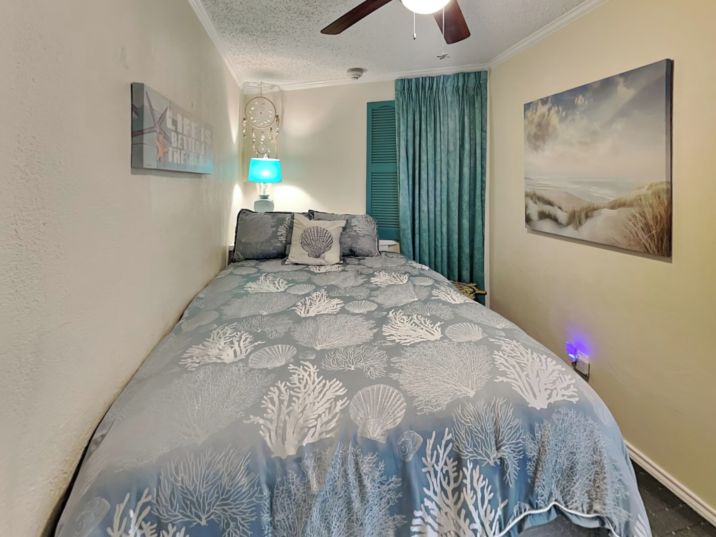 Un dormitorio con una cama, un ventilador de techo, una lámpara de pared, un cuadro y una ventana con cortinas.