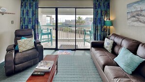A room with a brown leather sofa, a reclining chair, a wooden coffee table, and a view through a glass door to an outdoor area with chairs and a table. - Casa Del Mar 272-seashells & Tail Fins (Galveston)