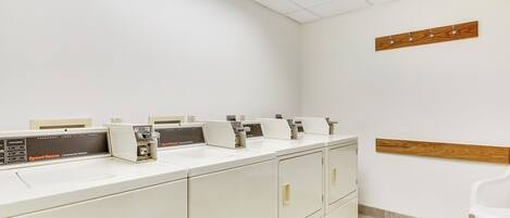 A commercial laundry area with multiple washing machines and dryers, a wooden shelf on the wall, and a white chair.