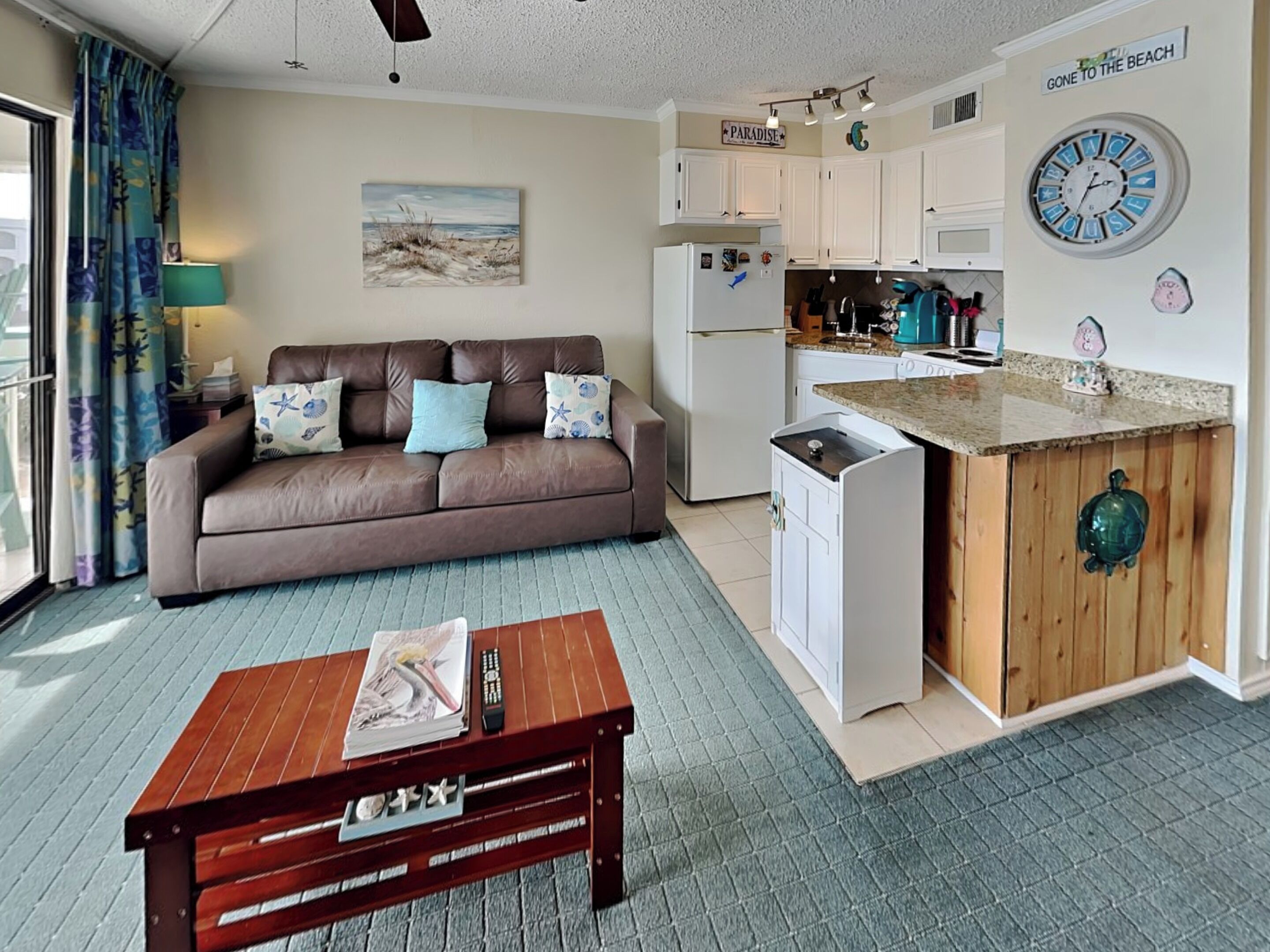 A compact living space with a kitchenette, a sofa with decorative pillows, a coffee table, and a view of the beach.