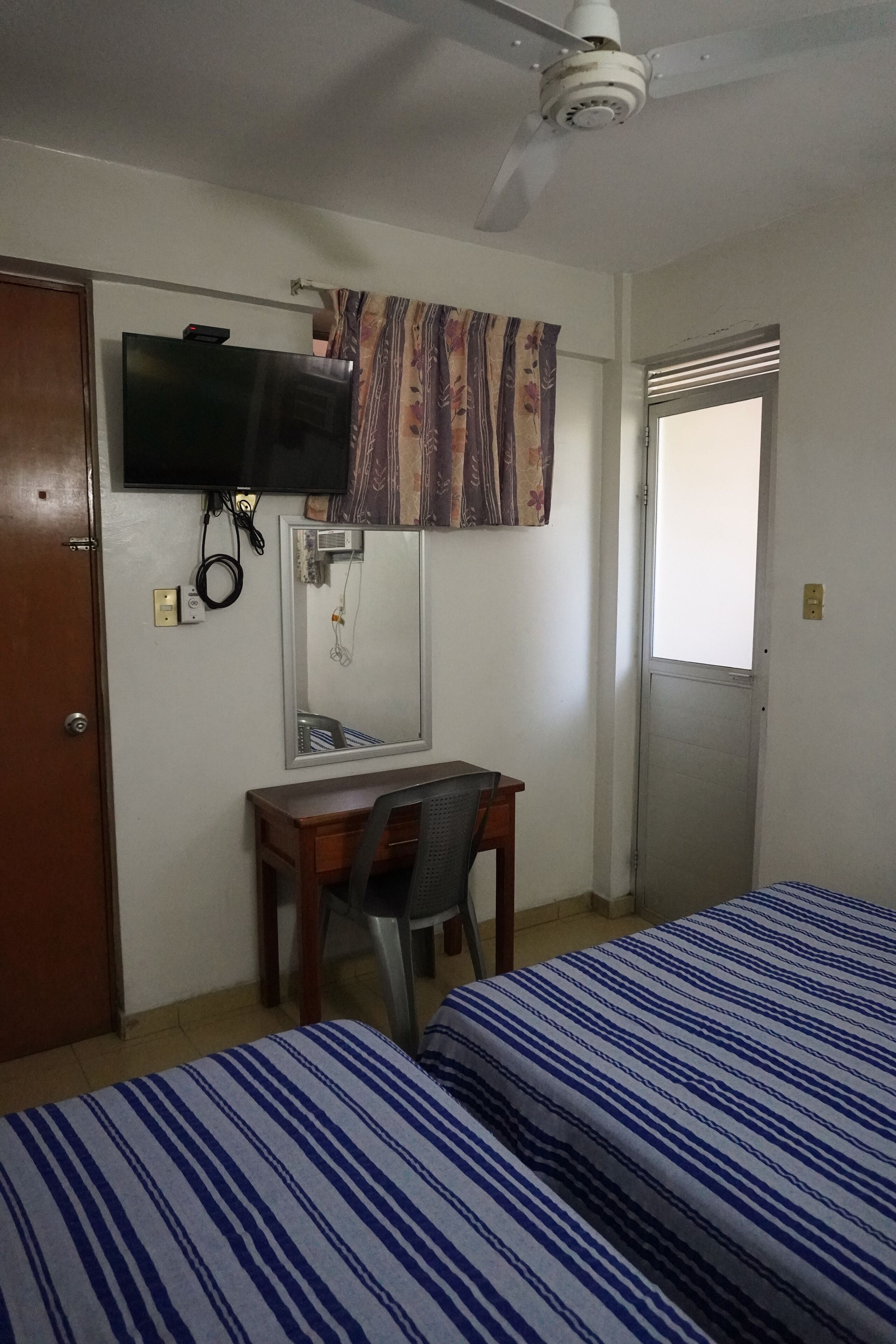 Habitación de hotel con dos camas, un escritorio con silla, televisión instalada en la pared, ventilador de techo y ventana con vistas.