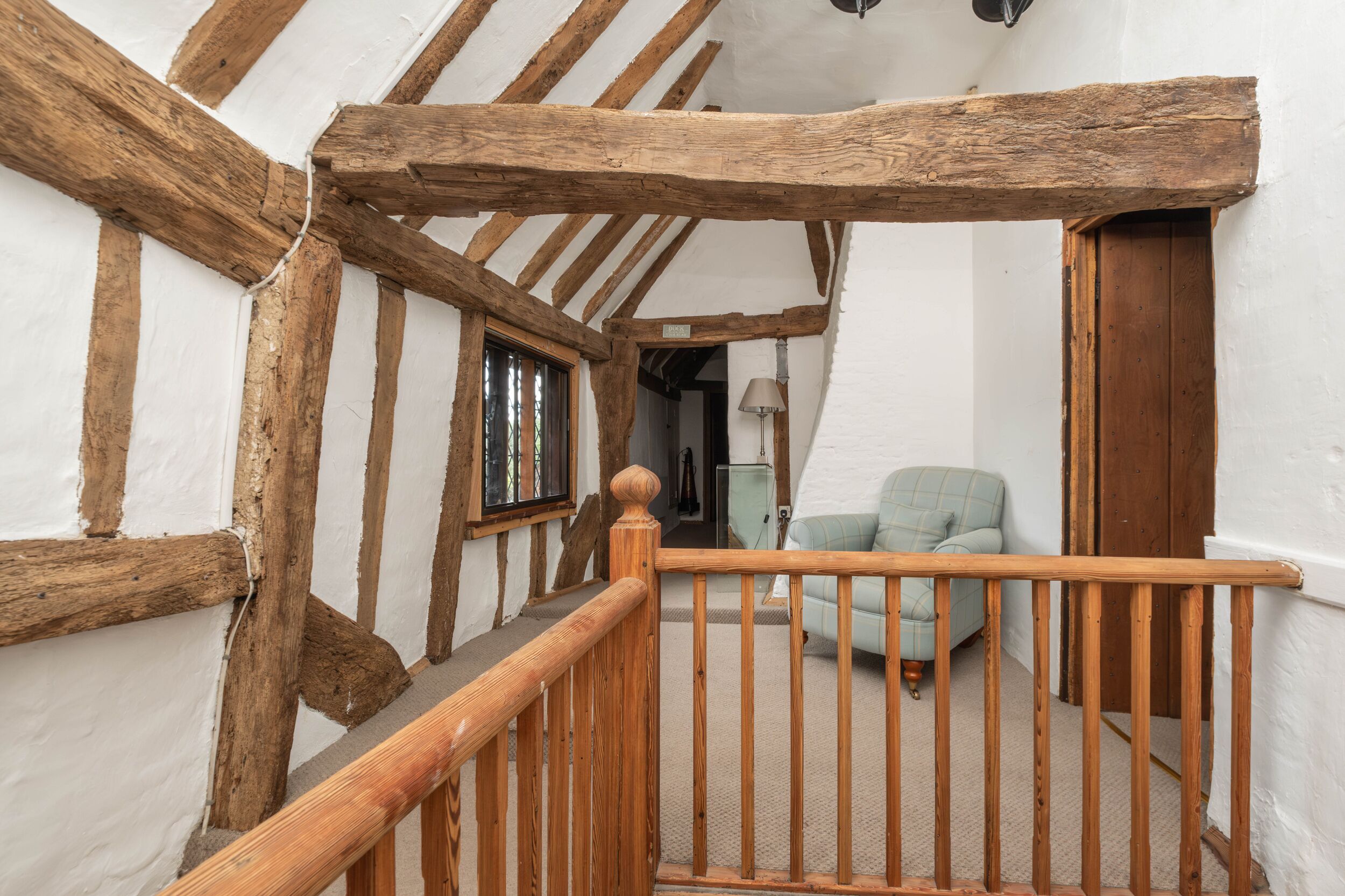A staircase with wooden beams and a wooden handrail leading to a room with a light blue armchair and a lamp.