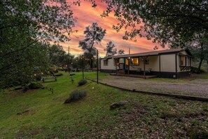 A rustic cabin with a porch, surrounded by a grassy area and trees, under a sunset sky. - Prvt Grounds: Cozy Yosemite Escape w/ Creek, Billiards, BBQ, Starlink, Pets ok (Mariposa)