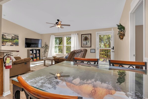 A spacious living area with a glass-top table, comfortable seating, a ceiling fan, and large windows offering a view of the outdoors. - Yosemite Nature Escape: Pvt Gem • Pets • BBQ • Cozy • Split BRs (Mariposa)