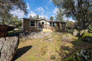 A single-story house with a front porch, surrounded by a stone retaining wall and large rocks. - Yosemite Nature Escape: Pvt Gem • Pets • BBQ • Cozy • Split BRs (Mariposa)