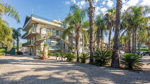 A multi-story building with balconies, palm trees, and a paved courtyard. - Apartment 'I Ciottoli Dello Ionio - Apt 10' with Sea View, Shared Terrace and Balcony (Mascali)