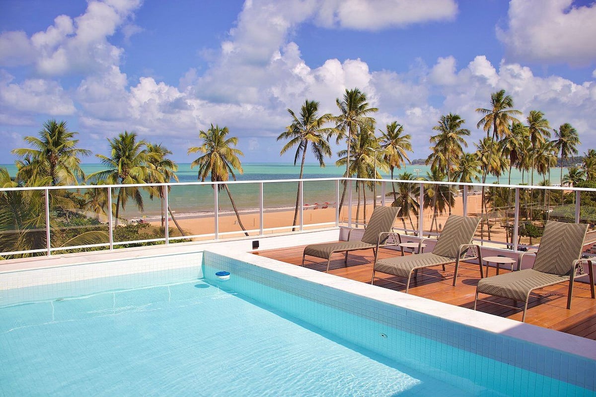 Ein Poolbereich mit Liegestühlen und Blick auf Strand und Palmen.