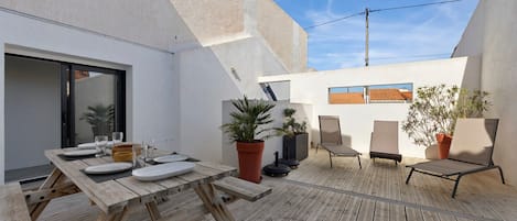 Terraza de madera con una mesa de comedor para cuatro personas, rodeada de macetas con plantas y dos sillones.