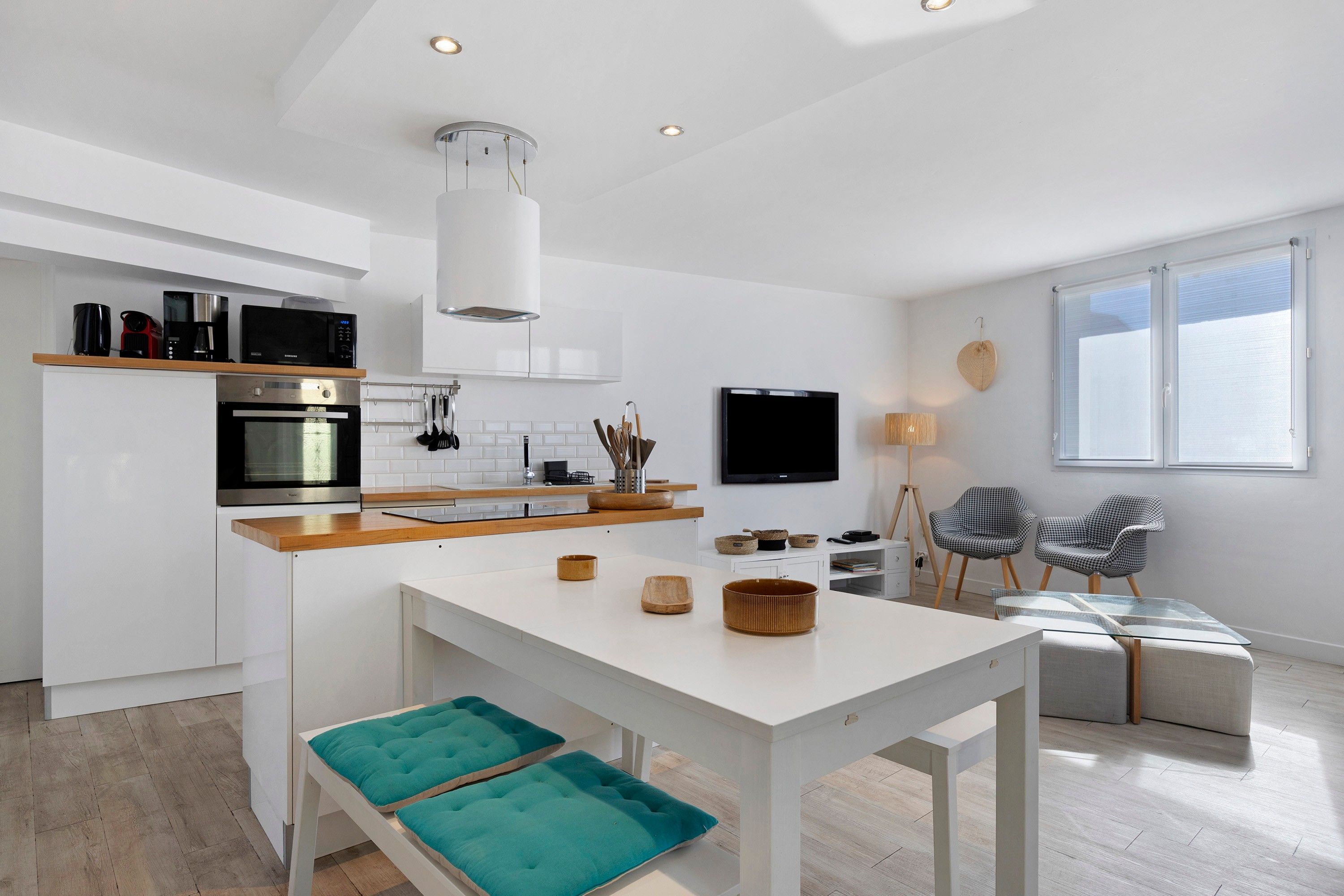 A modern kitchen with a central island, built-in appliances, and a dining area with a glass table and teal cushions.