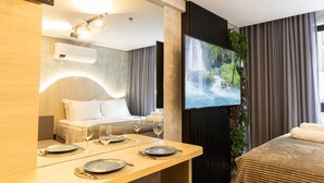Ein modernes Hotelzimmer mit einem Bett, einem an der Wand montierten Fernseher, einem Esstisch mit Tellern und Gläsern sowie einem großen Fenster mit Vorhängen.