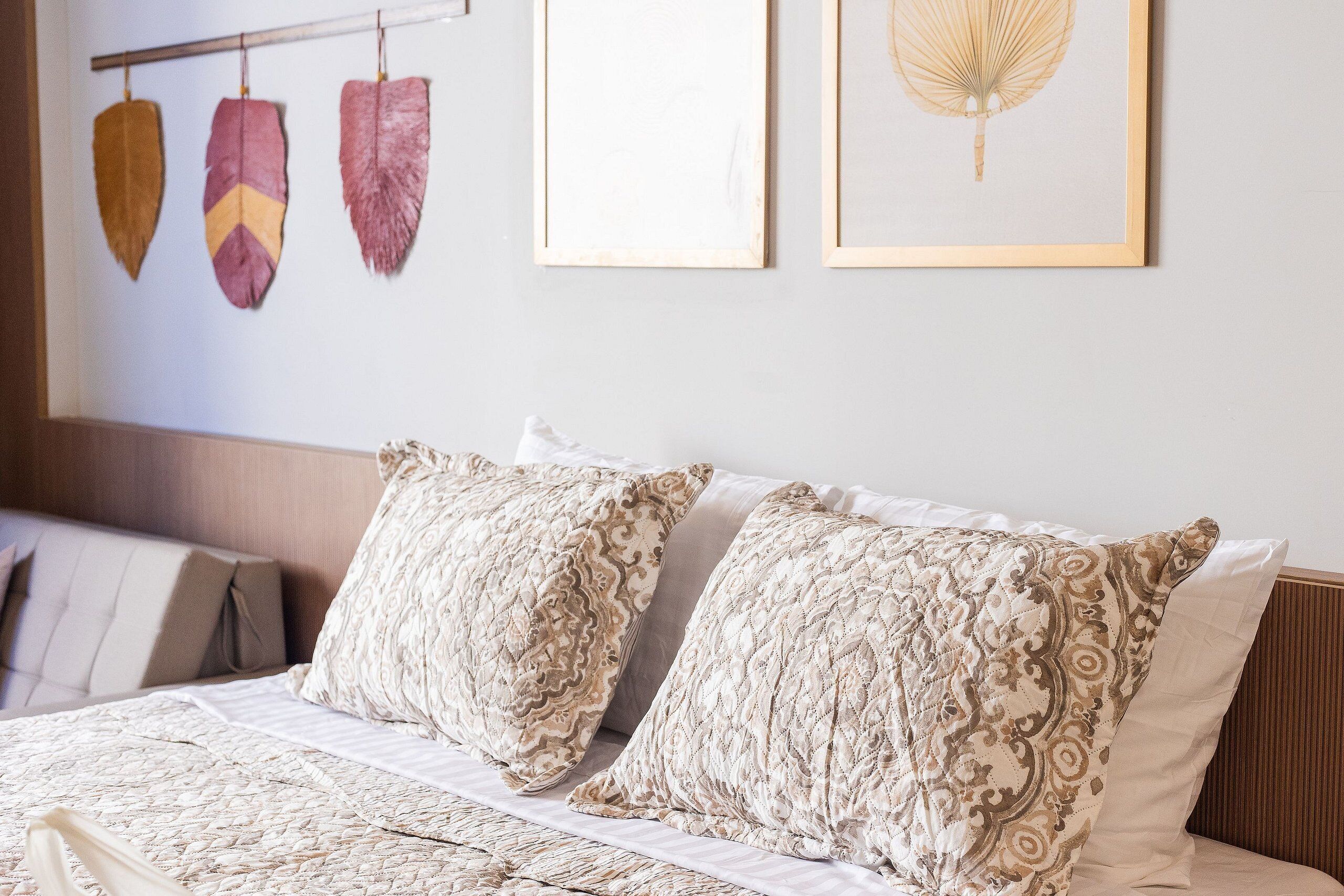 Una cama bien tendida con almohadones estampados, un cabecero y decoración en la pared, incluyendo una composición de plumas y una ilustración enmarcada.