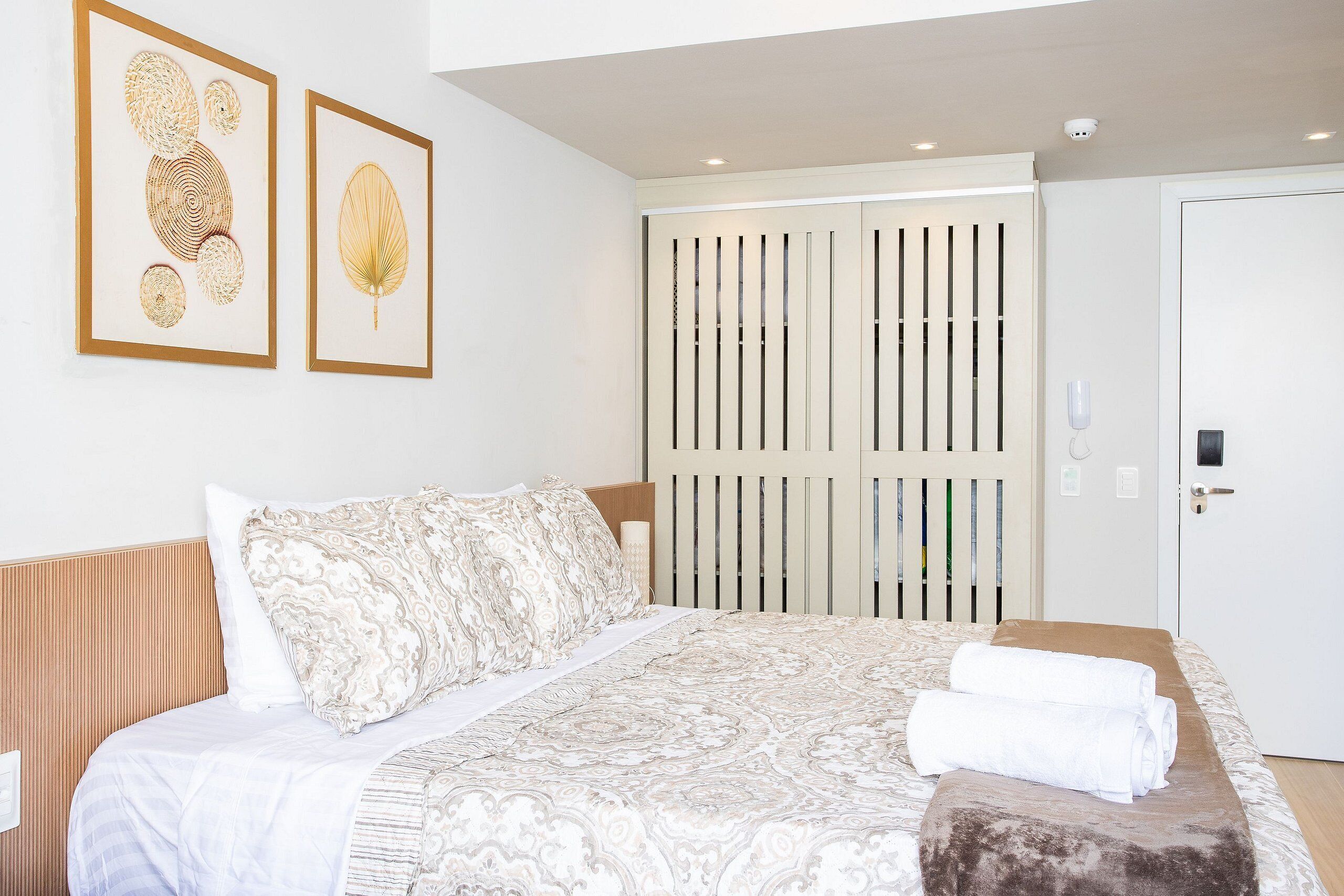A hotel room with a bed, pillows, a blanket, a towel, and two framed artworks on the wall.
