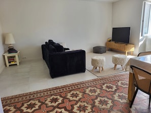 Ein Wohnzimmer mit einer schwarzen Couch, einem grauen Hocker, einem Fernseher auf einem Holzboden, einem kleinen Beistelltisch mit einer Lampe und einem gemusterten Teppich.