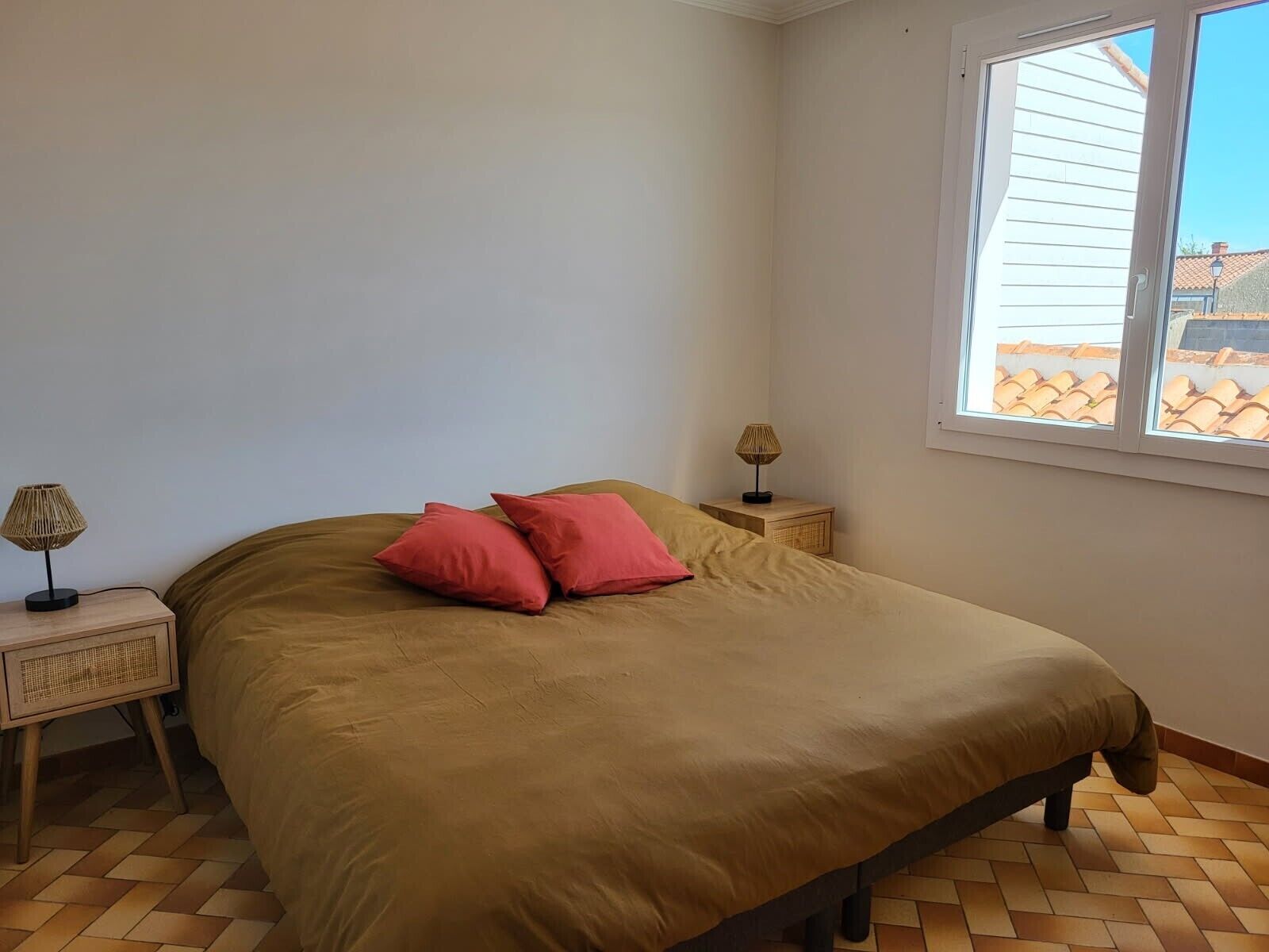 Un dormitorio con cama, dos mesitas de noche, una ventana y vistas a un tejado con tejas.