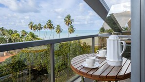 A balcony with a wooden table, a white coffee pot, and a cup, overlooking a beach with palm trees and the ocean. - Setai Yacht #522 - Beira mar by Carpediem (João Pessoa)