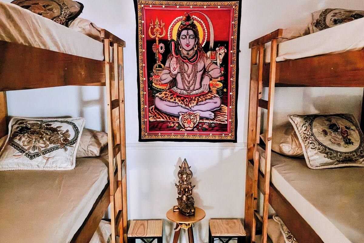 Una habitación con dos literas, una mesita con una estatua y un tapiz con una figura religiosa.