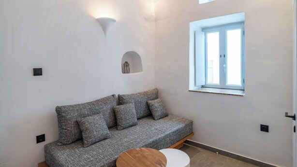 A modern living room with a grey sofa, a round wooden coffee table, and a small white table. There is a window with a view of the outside, and a wall-mounted light fixture. - Two story island apartment with quiet patio (Mykonos)