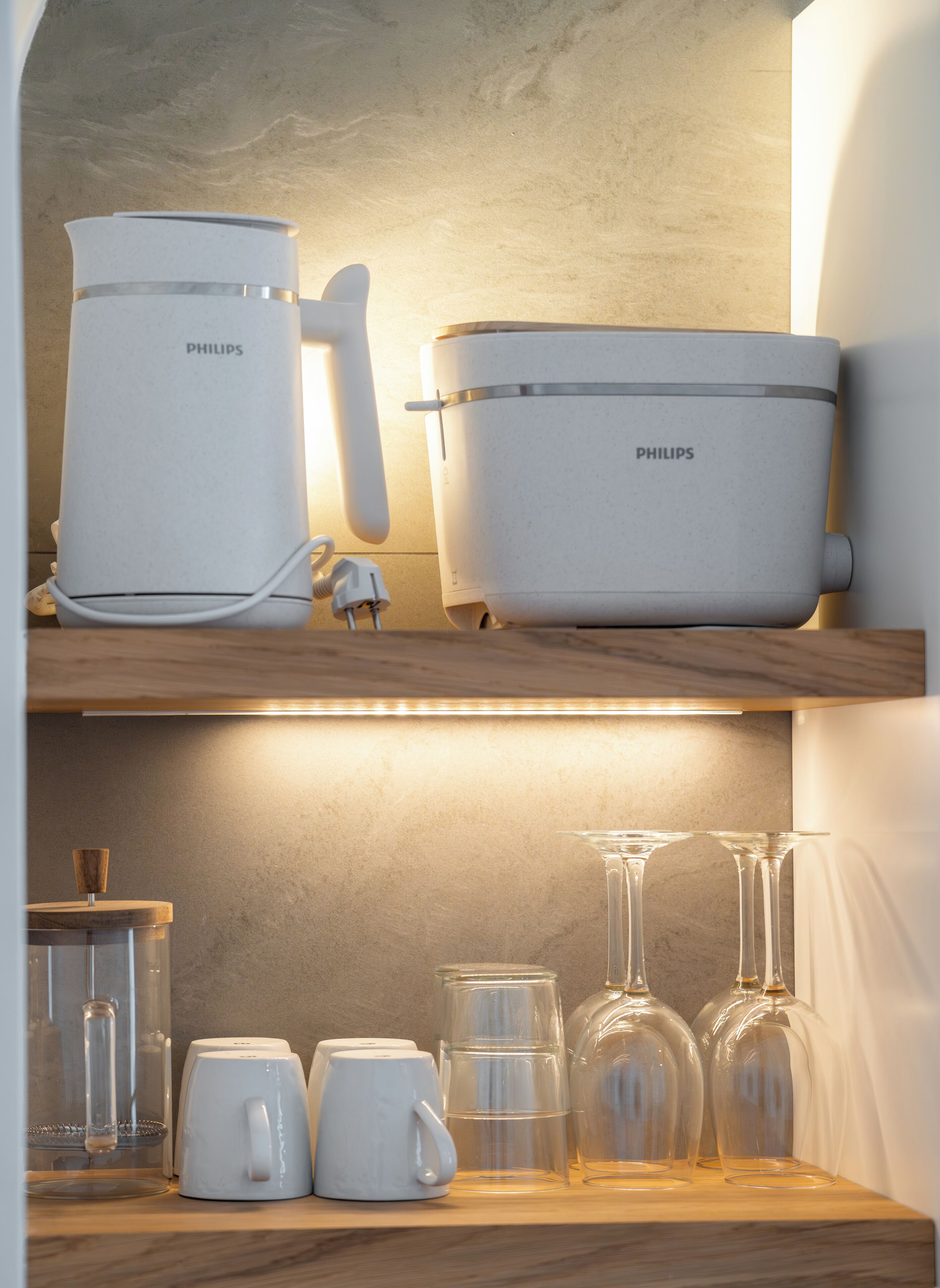 Un coin cuisine avec un appareil Philips posé sur le plan de travail, une étagère en bois au-dessus et divers objets en verre et ustensiles de cuisine en dessous.