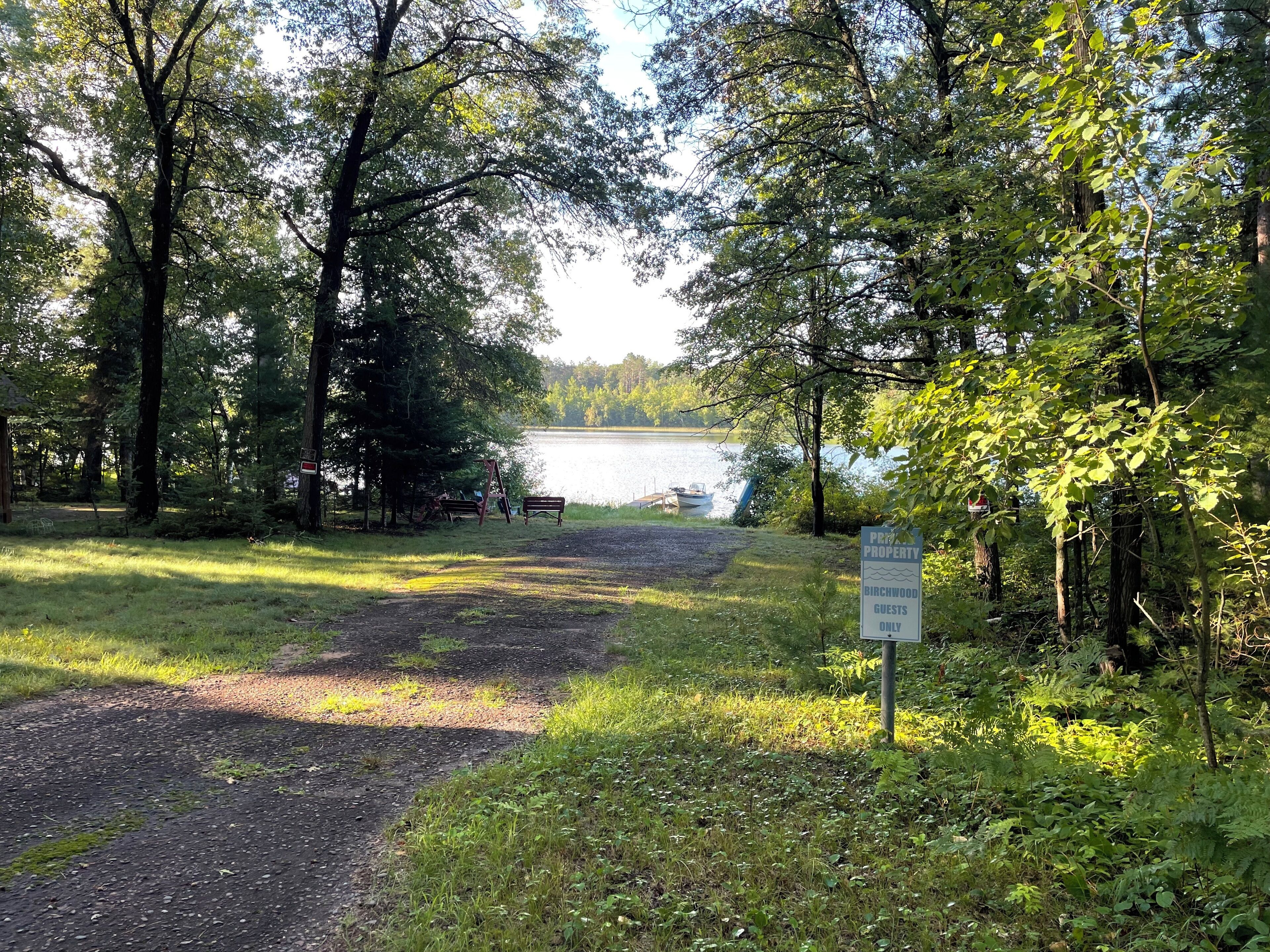 A wooded area with a gravel path leading to a lake, a bench, and a sign indicating private property for guests only.