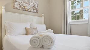 Un lit bien fait, avec du linge de lit blanc et des serviettes pliées, une tête de lit blanche et une fenêtre avec des rideaux.
