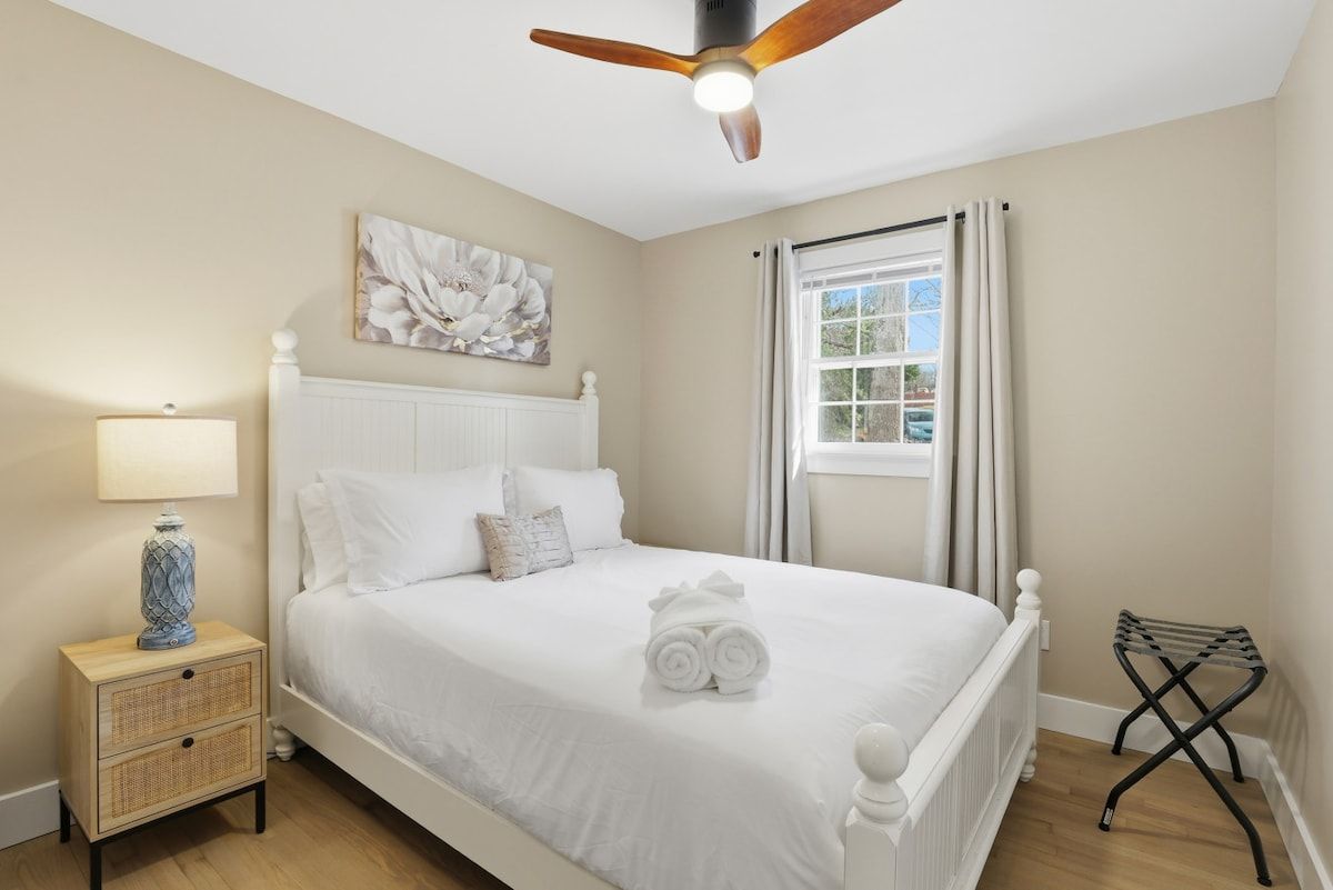 Une chambre à coucher comprenant un lit, une table de chevet, une lampe, un ventilateur de plafond, une fenêtre avec des rideaux et une œuvre d’art accrochée au mur.