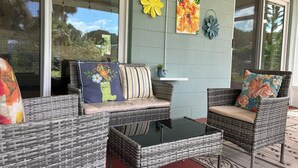 A covered outdoor seating area with wicker furniture, a glass table, and decorative wall art. - Three Blocks to Flagler - Fenced Backyard Pets OK (New Smyrna Beach)