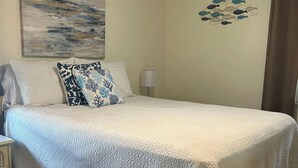 A neatly made bed with a white quilt, two pillows with a blue and white pattern, a wall art of fish, and a lamp on a bedside table. - Three Blocks to Flagler - Fenced Backyard Pets OK (New Smyrna Beach)