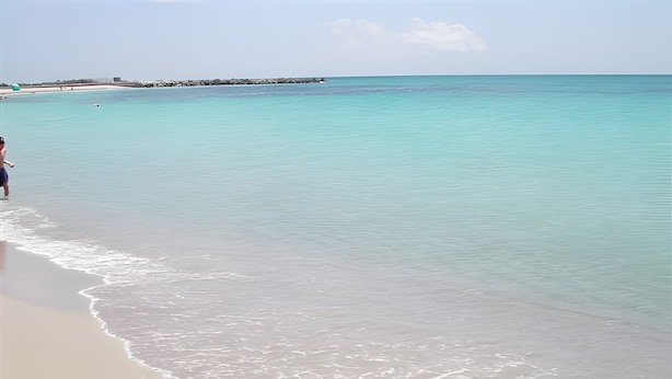 Une plage à l’eau turquoise et limpide, bordée d’un rivage sablonneux, avec, au loin, un promontoire rocheux.