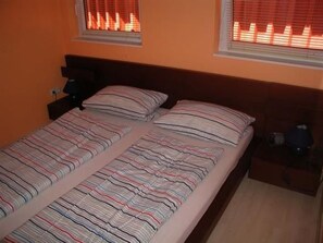 A double bed with striped bedding, two pillows, a nightstand with a lamp, and two windows with blinds. - Appartement in Spekendorf mit Terrasse by Interhome (Aurich)