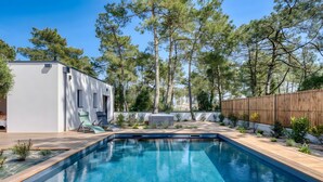 A swimming pool with a wooden deck, a white building with a covered patio, and a wooden fence. - La Villa des Pins d'Or – Family Villa with Pool and Spa in a Peaceful Natural Setting (La Tranche-sur-Mer)