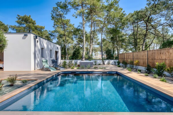 Une piscine avec une terrasse en bois, un bâtiment blanc doté d’un patio couvert et une clôture en bois.