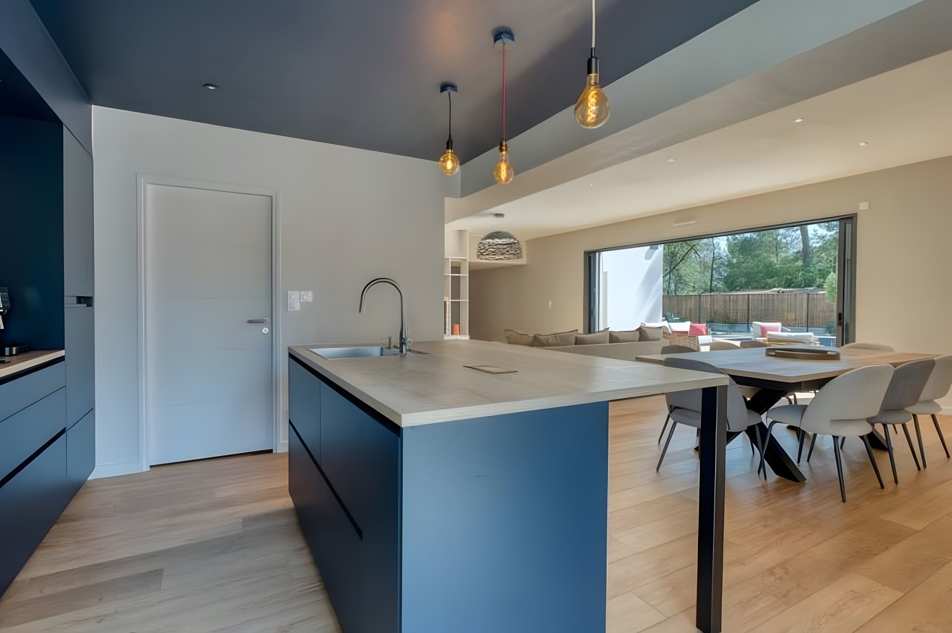 A modern kitchen with a central island, a dining area with a table and chairs, and a large window providing a view of the outdoors.
