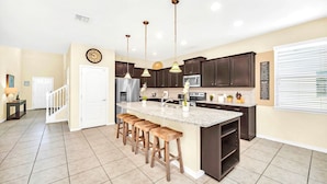 A modern kitchen with dark cabinets, a large island with stools, pendant lighting, and a clock on the wall. - 6BR Windsor Resort Home 15min Disney (Kissimmee)