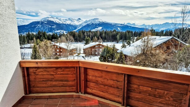 En balkon med trægelænder, der udover et sneklædt bjerglandskab og flere træbygninger.