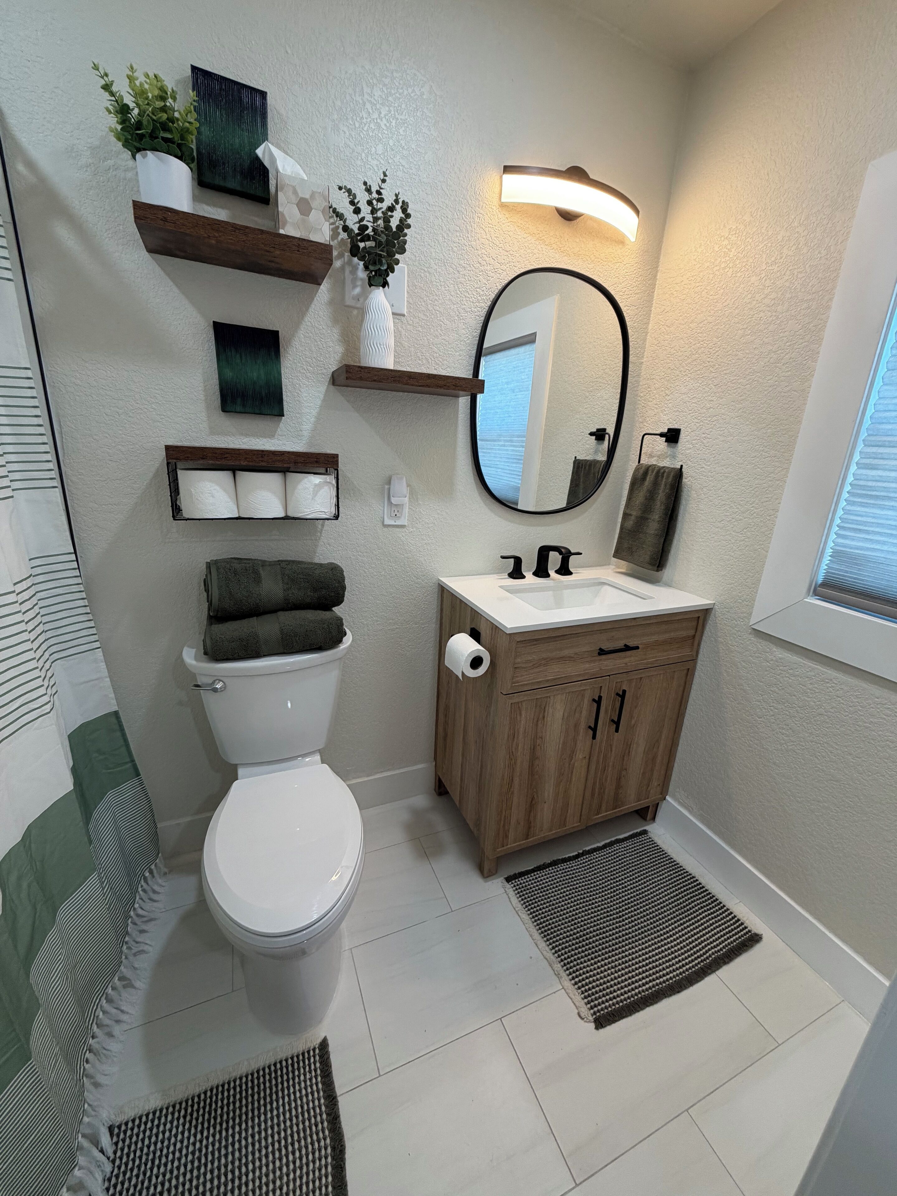 A modern bathroom with a toilet, sink, and mirror. There are shelves with plants and toiletries, a towel rack, and a window with blinds.