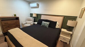 Un dormitorio ordenado con cama, una silla, una mesita de noche, lámpara, cómoda y aire acondicionado.