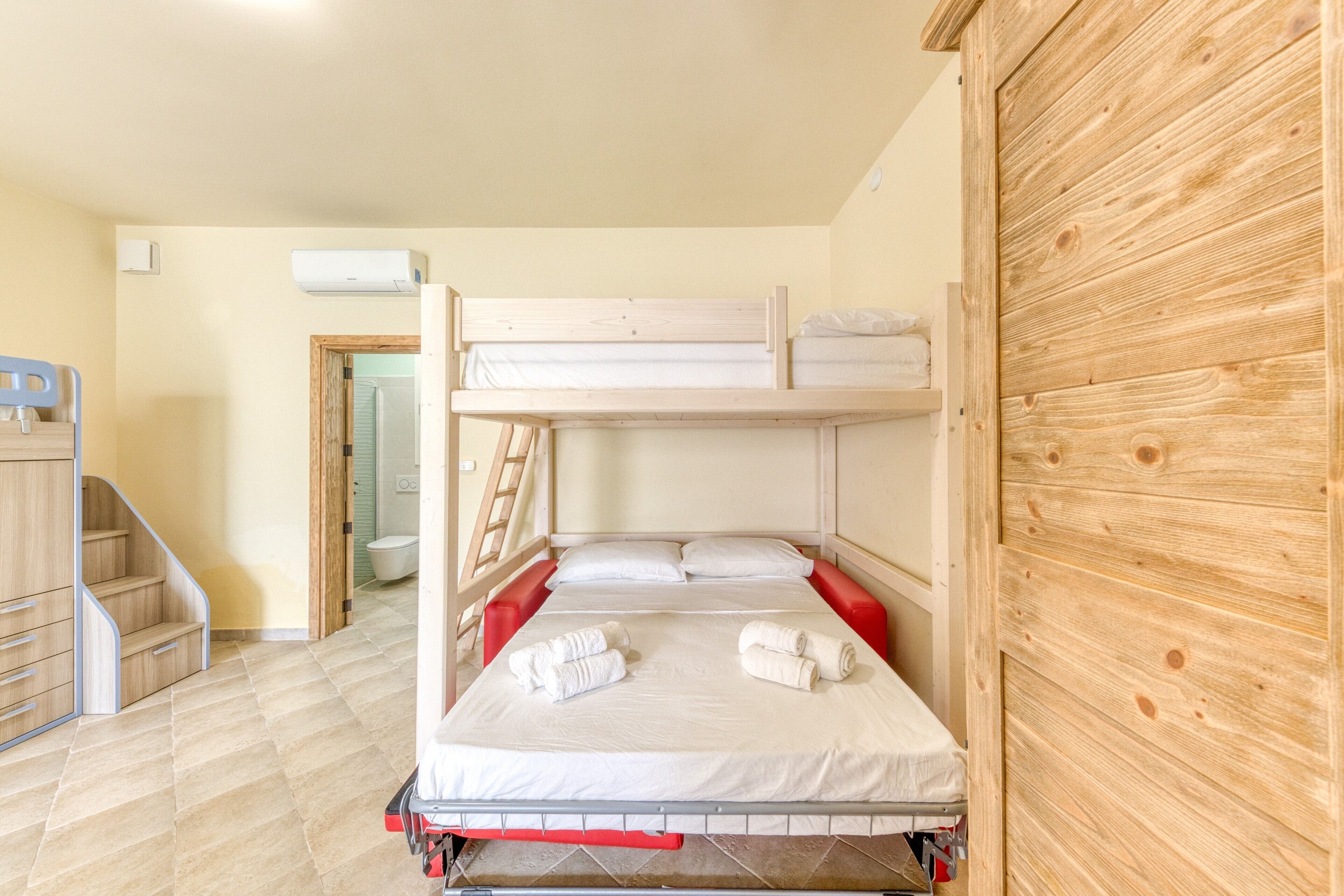 Ein Schlafraum mit Etagenbetten, einem Holzschrank, einem Schreibtisch und einem Badezimmer im Hintergrund.