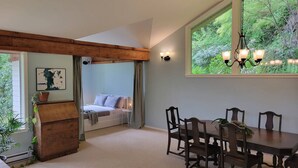 A spacious room with a large dining table, a cozy seating area with a sofa, and a view of greenery outside. - Relaxing 1 bdr + den rainforest retreat with patio (Bowen Island)