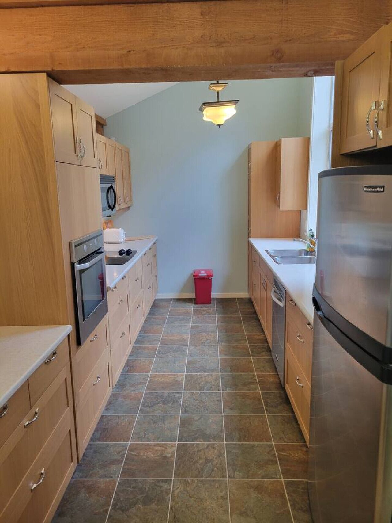 Eine schmale Küche mit Holzschränken, einem Edelstahlkühlschrank, einer Mikrowelle und einem roten Mülleimer.