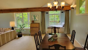 A dining area with a long wooden table, six chairs, a chandelier, and a view of greenery through large windows. - Relaxing 1 bdr + den rainforest retreat with patio (Bowen Island)