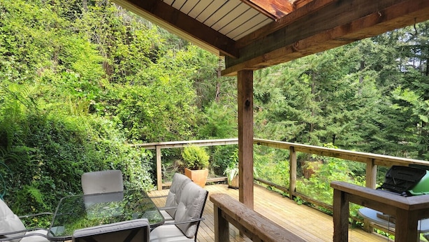 A wooden deck with outdoor furniture, including chairs and a table, surrounded by lush greenery. - Relaxing 1 bdr + den rainforest retreat with patio (Bowen Island)