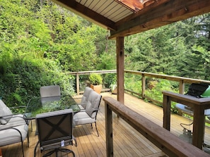 Una terraza de madera con mobiliario exterior, incluyendo sillas y una mesa, rodeada de abundante vegetación.