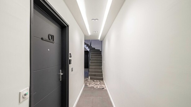Ein enger Flur mit einer schwarzen Tür mit der Aufschrift "01" und einer Treppe im Hintergrund.