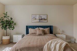 A bedroom with a bed, a nightstand, a lamp, a plant, and a painting on the wall.