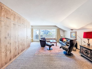 A spacious living room with a large wooden wardrobe, a black leather sofa, a red lamp, and a view of the city through a large window. - Matten (Utoring) by Interhome (Zermatt)