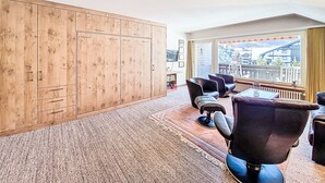 Ein Hotelzimmer mit großem Kleiderschrank, einer Sitzecke mit Stühlen, einem kleinen Tisch und einem großen Fenster mit Blick auf Gebäude und einen Balkon.